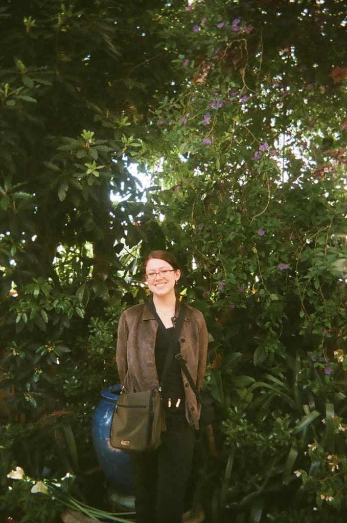 An image of myself in front of trees. A white woman with dark red hair, glasses, black clothes and a brown suede jacket on with my camera bag and a green messenger bag.